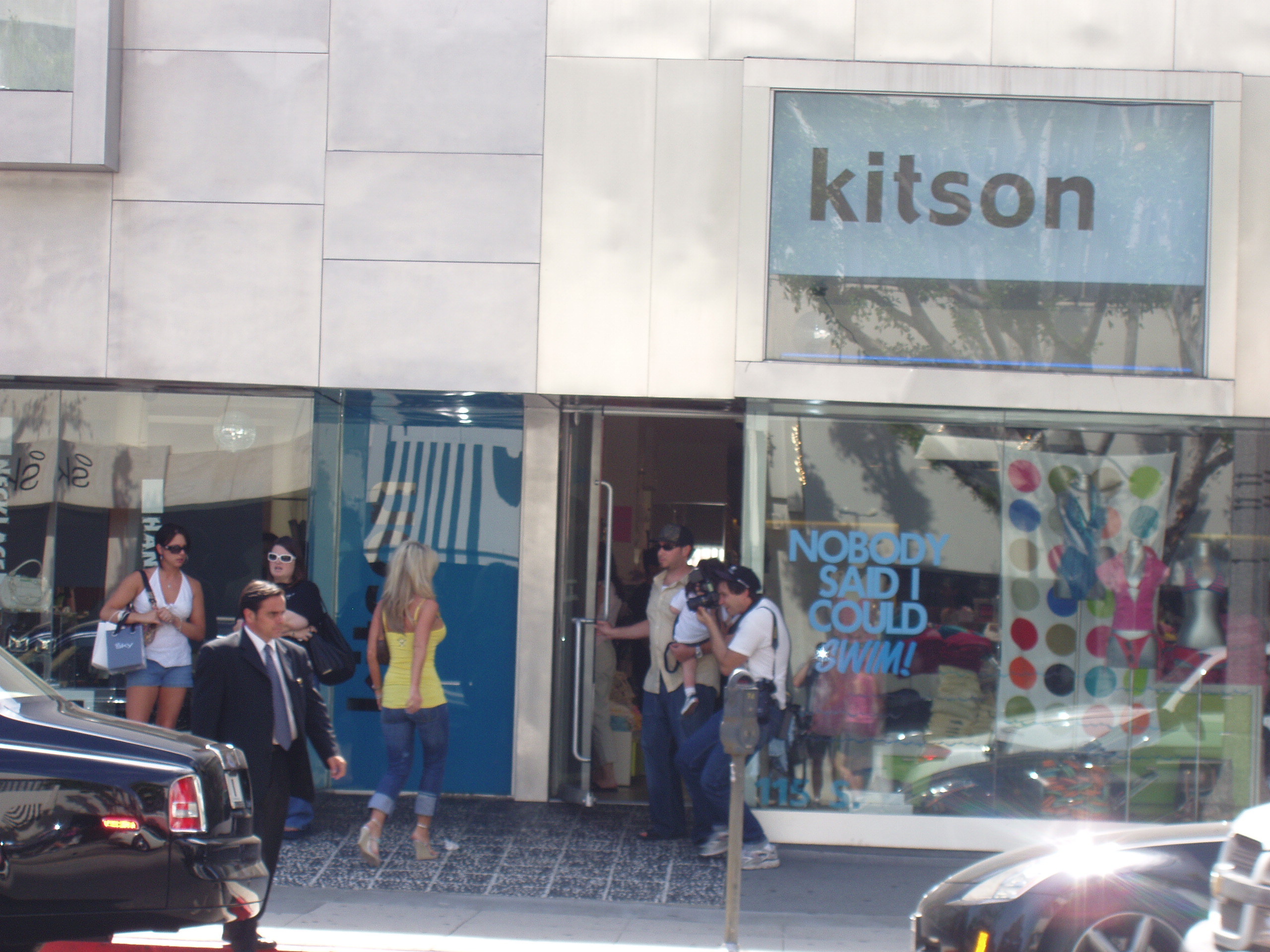 Paparazzi take photos as a blond woman walks into a store. A limo and security person or driver is in the foreground.